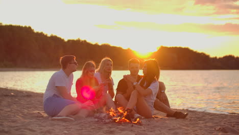 Two-men-and-three-young-female-student-are-sitting-around-bonfire-on-the-beach-with-beer.-One-of-the-female-students-is-telling-interesting-story.-Another-one-is-checking-her-mobile-phone-at-sunset-in-summer-evening-on-the-lake-coast.